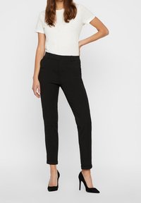 Femme se tenant debout portant un haut à manches courtes blanc, un pantalon noir slim-fit jusqu'à la cheville, et des chaussures à talons hauts noires sur un fond uni.