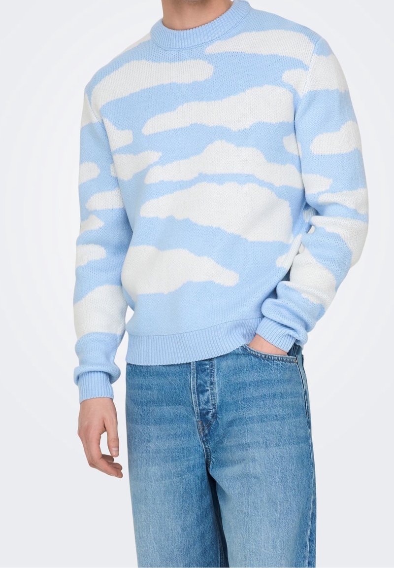 Homme portant un pull bleu clair avec des motifs de nuages blancs et un jean bleu, une main dans la poche, devant un fond uni.