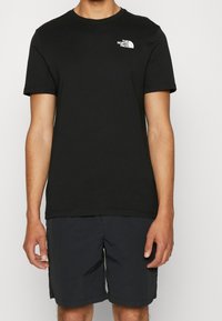 Black cotton t-shirt with a round neckline and short sleeves. Features a small white logo on the chest. Paired with dark shorts.