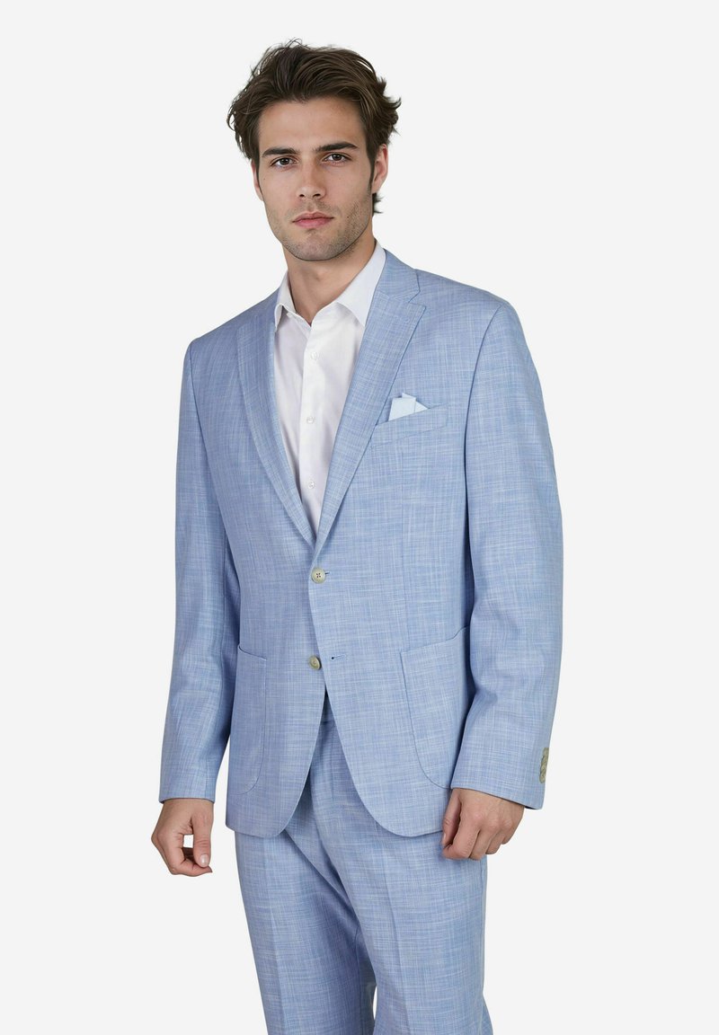 Jonge man in een lichtblauw kostuum met een wit shirt en een pochetta, staande tegen een eenvoudig witte achtergrond.