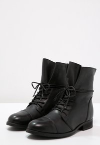 Botas de tobillo de cuero negro con puntera redondeada, detalle de puntera con costura, cordones en la parte frontal y un eje ligeramente ajustado. Suela plana y acabado suave.