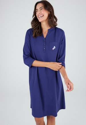 Mujer sonriente, vestida con un vestido azul marino hasta la rodilla, con mangas tres cuartos y un pequeño diseño floral en el bolsillo del pecho.