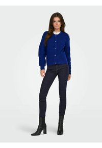 Modro pletena jopica z dolgimi rokavi, z gumbi, z rebrastimi manšetami, nošena preko bele srajce s ovratnikom, v kombinaciji s temnimi ozkimi kavbojkami in črnimi škornji.