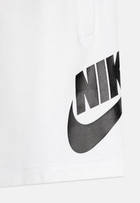 Beli kratke hlače iz mehkega materiala, ki imajo velik črn Nike logo in obliko klina, z gladko teksturo in čistim šivom.