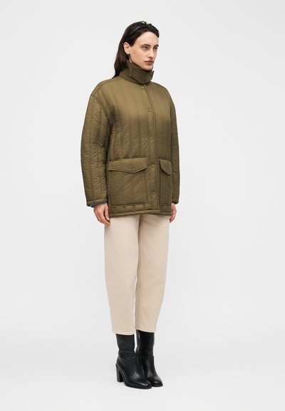 Γυναίκα που φοράει πράσινο λαδί καπιτονέ μπουφάν με ψηλό γιακά, μπεζ παντελόνι και μαύρα μπότες με τακούνι, στέκεται μπροστά σε απλό φόντο.