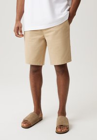 Beżowe bawełniane szorty o prostym kroju, z bocznymi kieszeniami i podwiniętymi brzegami. Dobrane do nich taupe sandały wsuwane, wykonane z zamszu.