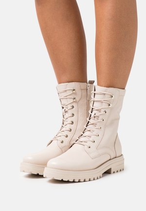 Stivali in pelle beige alla caviglia con lacci e suole robuste, indossati su gambe inferiori, fotografati su uno sfondo bianco semplice.