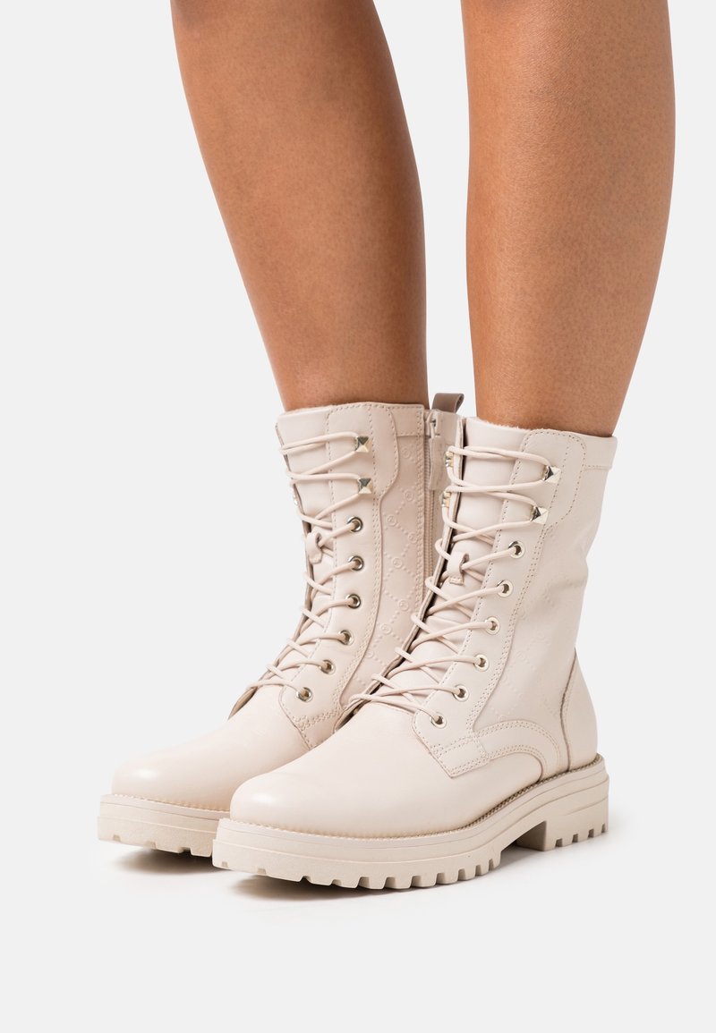 Beiga snörade läderankelstövlar med robusta sulor på personens underben, fotograferade mot en enfärgad vit bakgrund.