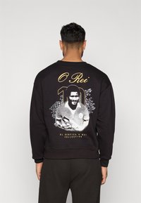Felpa nera con scollo rotondo, decorata con una grafica di un calciatore e la scritta "O Rei 10" in oro. Maniche e bordo inferiori a costine.