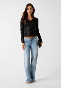 Black ribbed long-sleeve top with button details paired with light blue wide-leg jeans, and black clutch. Model wearing open-toed sandals.