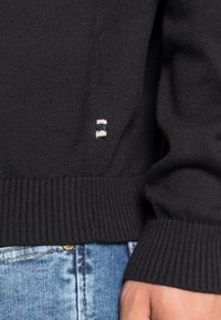Sort strikket sweater med ribbet manchetter, prydet med et lille stoflogo med rød, hvid og blå accent på den nederste side.