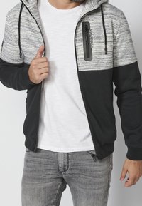 Felpa nera e grigia con zip, dotata di una sezione superiore testurizzata, tasca sul petto con zip e lacci regolabili sul cappuccio.