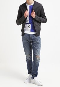 Schwarze Zip-Jacke über einem weißen T-Shirt mit blauem Grafik, kombiniert mit hellblauen, zerrissenen Jeans und weißen Sneakers.
