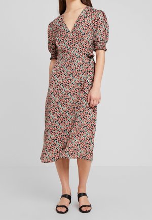 Donna che indossa un vestito avvolgente floreale con maniche a sbuffo e infradito nere, in piedi davanti a uno sfondo grigio chiaro uniforme.