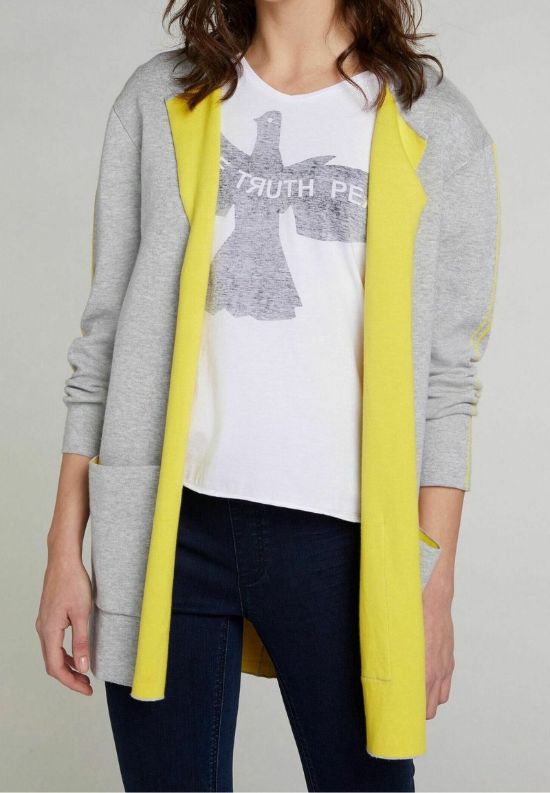 Grå cardigan med gula detaljer, mjukt tyg, långa ärmar, öppen fram, två fickor. Bärs över en vit t-shirt med en fågelgrafik.