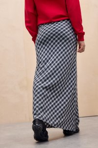 Egy személy piros hosszúujjú felsőt, fekete-fehér kockás hosszú szoknyát és fekete texturált talpú cipőt visel, és egy szürke padlón áll.