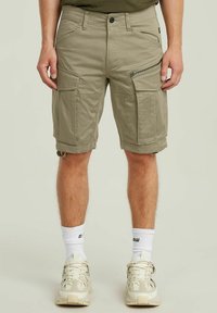 Pantaloni cargo beige con più tasche, inclusa una tasca laterale con zip, realizzati in un tessuto resistente. Presentano risvolti e passanti per la cintura.