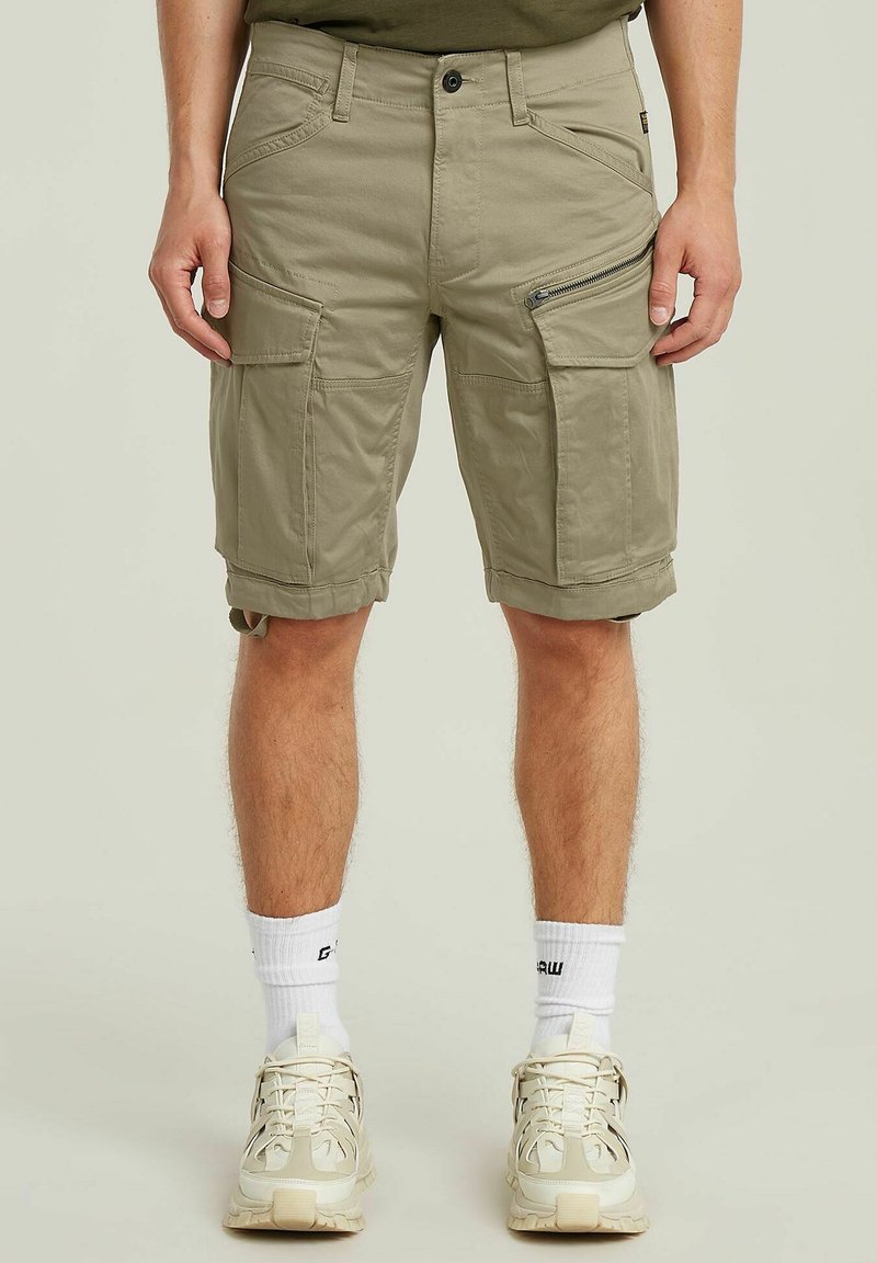 Pantaloni cargo beige con più tasche, inclusa una tasca laterale con zip, realizzati in un tessuto resistente. Presentano risvolti e passanti per la cintura.