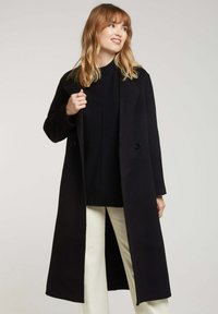 Manteau en laine noir à coupe longue et droite, à double boutonnage, avec des revers crantés, porté par-dessus un pull foncé et un pantalon crème.