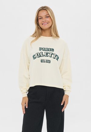 Crème sweatshirt met de tekst "PARIS COLETTE CLUB" in donkergroen. Heeft een relaxte pasvorm, lange mouwen en een cropped zoom. Gemaakt met zwarte broek.