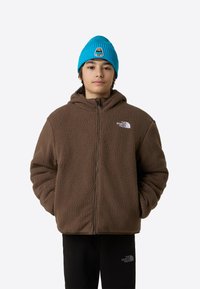 Rjava jopica iz flisa z zadrgo in kapuco ter teksturirano površino. Kombinirana z svetlo modro rebrasto kapo z grafično nalepko.