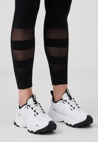 Leggings noirs avec des panneaux transparents rayés, associés à des baskets blanches et noires Nike avec une semelle texturée et un design à lacets.