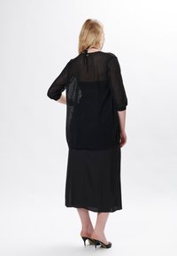 Schwarzes Netzoberteil mit Dreiviertelärmeln, übereinander getragen mit einem einfarbigen schwarzen Rock, der ein Rückenknoten-Detail aufweist. Durchscheinende Textur über das gesamte Outfit.