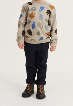 Kind trägt beigen Pullover mit bunten Monster-Mustern, dunkle hochgekrempelte Hose und grüne Schnürstiefel, steht vor einfachem Hintergrund.