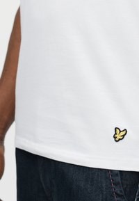 Άτομο που φοράει λευκό μπλουζάκι με ένα μικρό κίτρινο σύμβολο πουλιού κοντά στο τελείωμα και σκούρα μπλε τζιν με ροζ ραφές.