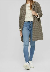 Abrigo a cuadros beige y marrón sobre un suéter de punto blanco, combinado con jeans ajustados azules y zapatillas blancas.