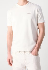 Muž oblečený v jednoduchém bílém tričku s krátkým rukávem značky Calvin Klein a bílých kalhotách, s jednou rukou v kapse.
