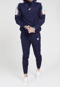 Marineblauw trainingspak van zachte stof, met een capuchontrui met een gekleurd streepdesign en slim-fit broek. Witte sneakers.
