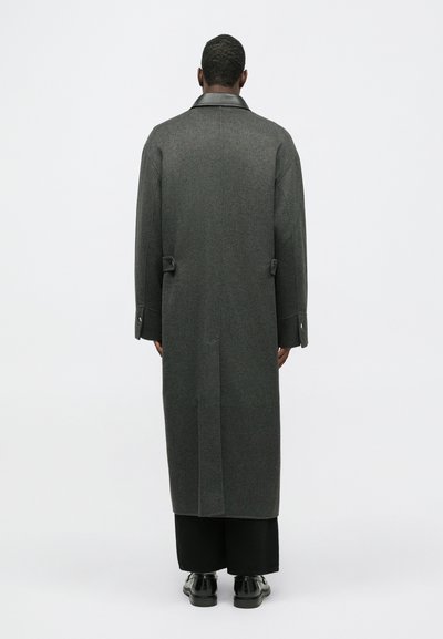 Garšs pelēks vilnas mētelis ar augstu apkakli, aizmugurējo venti un pogu akcentiem. Gluda tekstūra un minimālisma dizains. Valkāts virs melnajām biksēm.