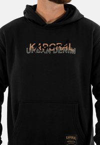 Sudadera negra con bolsillo frontal, que presenta el texto gráfico "KAPORAL URBAN DENIM" en colores contrastantes. El texto incluye estilos en bloque y cursiva.
