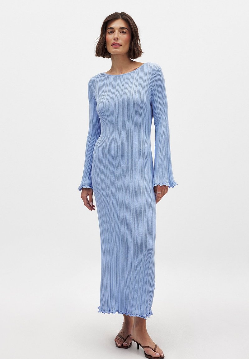 Pitkä, vaaleansininen neulemekko, jossa on ribbitekstuuria, pyöreä pääntie, levenevät hihat ja aaltoileva helma. Jalkineissa on ohuet hihnat.