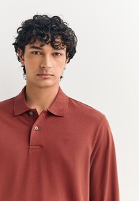 Camiseta tipo polo color óxido con cuello, dos botones y un pequeño emblema bordado en el pecho. Tejido suave y texturizado.