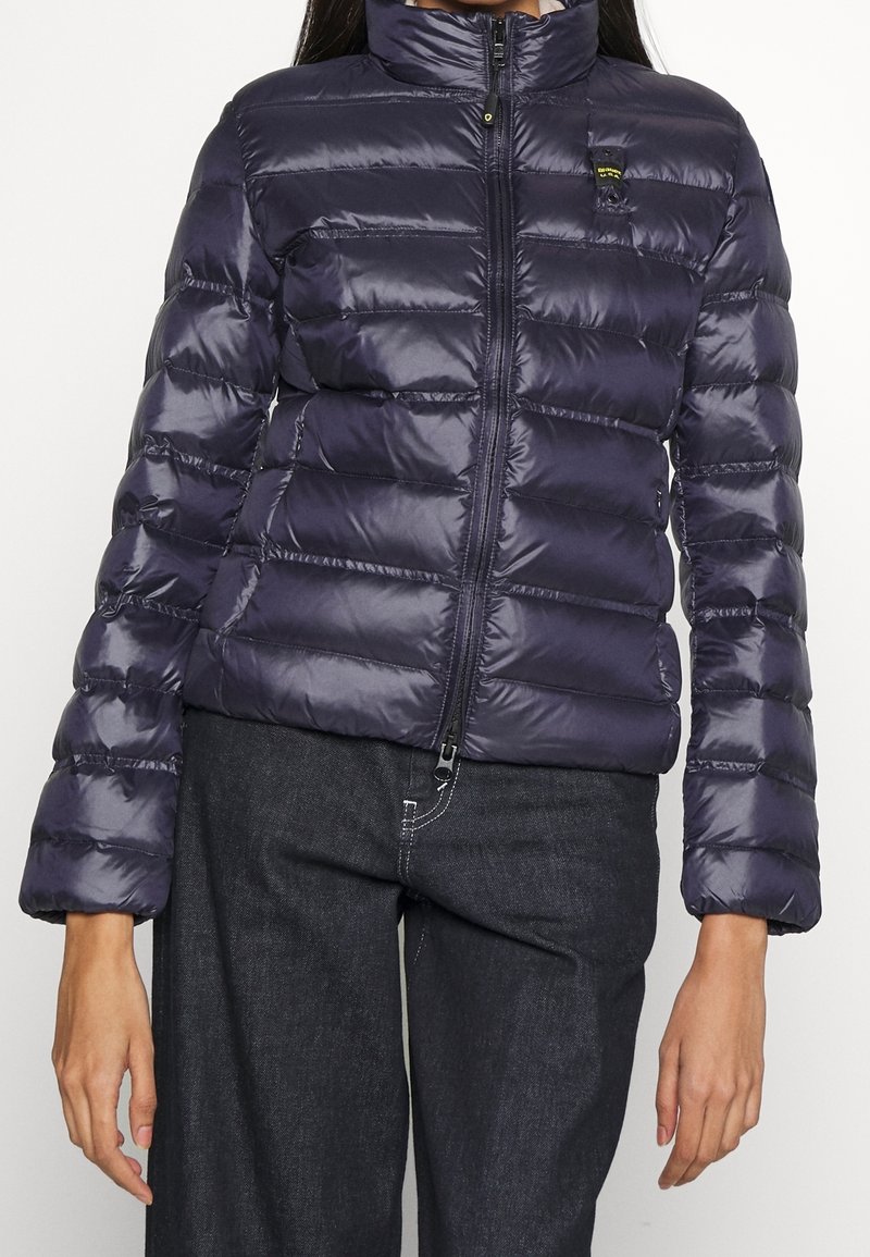 Femme portant une doudoune matelassée bleu marine avec fermeture éclair devant et un jean en denim foncé, debout devant un fond blanc uni.