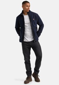 Marine fleecejakke med høj krave, lynlås og lommer foran, parret med en hvid T-shirt og mørke jeans, samt brune støvler.