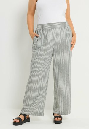 Erwachsener trägt hoch taillierte, weit geschnittene graue Hose mit weißen vertikalen Streifen und schwarze Plattform-Sandalen, Hände in den Taschen.