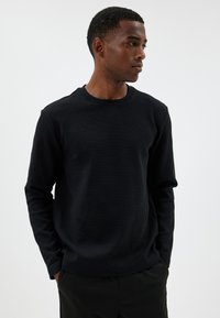 Schwarzes, geripptes Langarmshirt mit Rundhalsausschnitt, das aus strukturiertem Stoff gefertigt ist und eine lässige Passform hat, getragen über schwarzen Hosen.