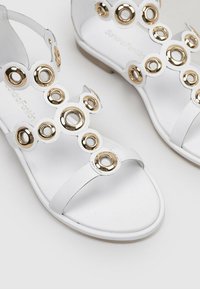 Sandalias blancas con acentos circulares dorados en las correas, con suela plana y un diseño minimalista, fabricadas con material sintético liso.