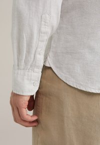 Camisa ligera a rayas en crema y gris claro con mangas con puños que presentan dos botones blancos; textura suave y corte relajado.
