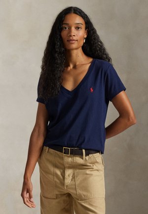 Γυναίκα με μακριά σγουρά μαλλιά, φοράει μπλε σκούρο T-shirt με V-λαιμόκοψη και κόκκινο λογότυπο και μπεζ παντελόνια με μαύρη ζώνη, να στέκεται μπροστά σε ουδέτερο φόντο.