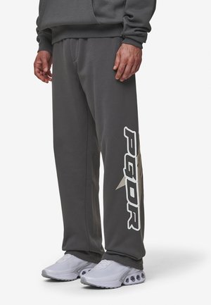 Mężczyzna w ciemnoszarych spodniach dresowych z napisem "PEGOR" i grafiką gwiazdy na lewej nogawce, w białych sportowych butach.