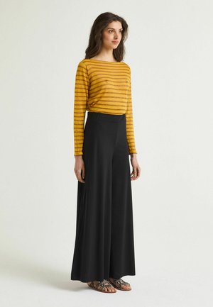 Donna in piedi con maglia a maniche lunghe a righe gialle e marroni, pantaloni neri a gamba larga e sandali bassi con cinturini su uno sfondo semplice.