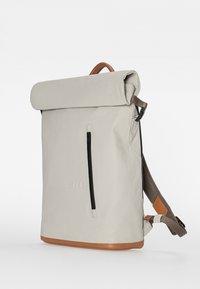 Lichtgrijze rugzak met oprolbare bovenkant en een gladde textuur, voorzien van een voorvak met ritssluiting en een bruine leren bodem en accenten. Verstelbare bandjes.