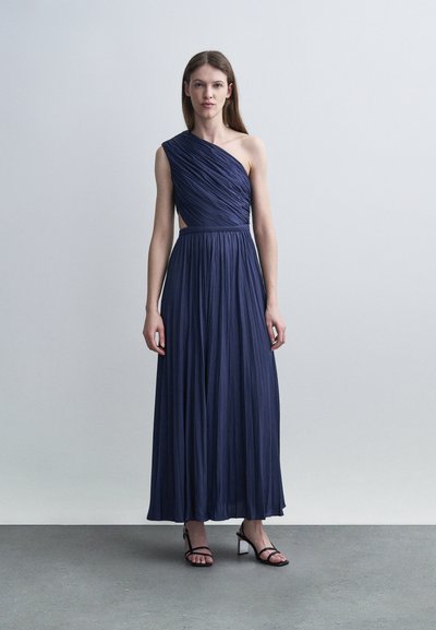 Femme debout portant une robe maxi plissée bleu marine à une épaule et des sandales noires à talons avec brides, sur un sol gris devant un mur gris clair.