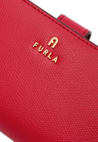 Portefeuille en cuir rouge avec une surface texturée, un logo en métal doré et une fermeture à agrafe. Design compact avec des bords lisses.