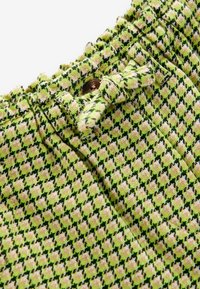 Struktureret grøn og gul mønstret stof med en samlet talje og dekorativ snørering, der har et mørkt houndstooth-mønster. Bronze knapdetail.