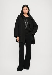 Zwarte oversized jas met drukknoopjes, hoge kraag en een katoenen stofmix. Gepaard met een zwart graphic T-shirt en wijde zwarte broeken.